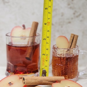 May include: Two glasses of a reddish-brown beverage, each with a cinnamon stick and apple slice. A yellow tape measure shows the taller glass is about 5 inches high. Sliced apples and cinnamon sticks are in the foreground.