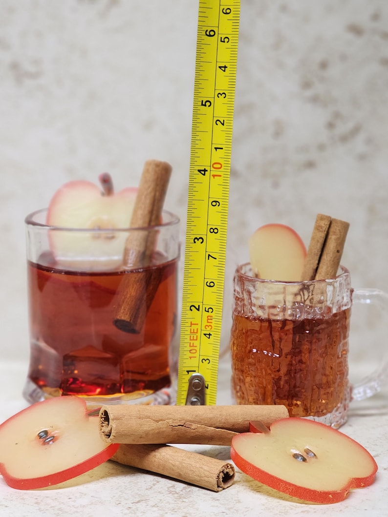 May include: Two glasses of a reddish-brown beverage, each with a cinnamon stick and apple slice. A yellow tape measure shows the taller glass is about 5 inches high. Sliced apples and cinnamon sticks are in the foreground.