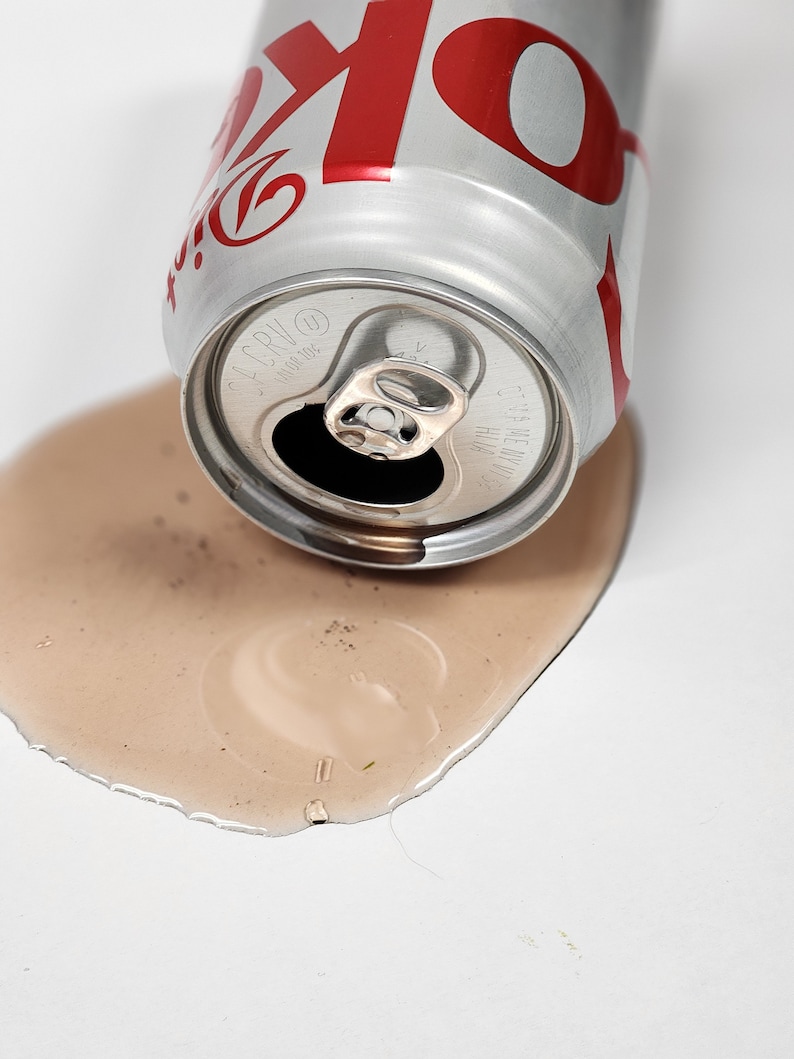 May include: A silver can with a red "Coke" logo is lying on its side on a white surface. The can is leaking a brown liquid.