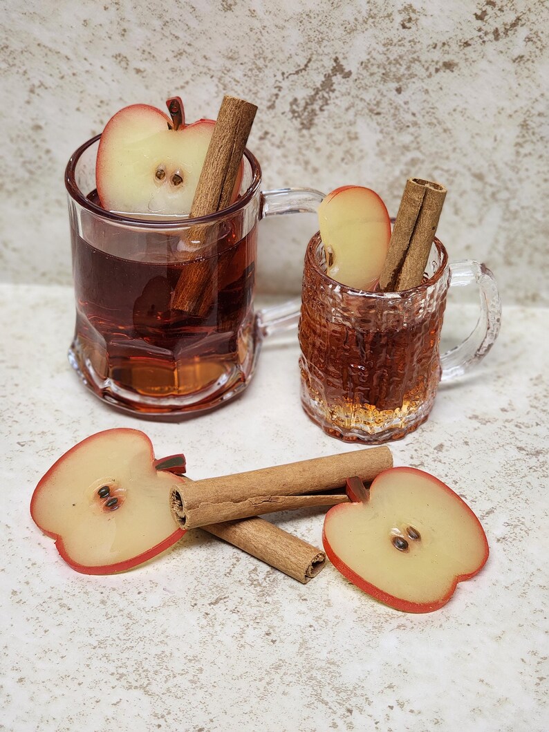 May include: Two clear glass mugs filled with a reddish-brown liquid, each containing an apple slice and a cinnamon stick. Two additional apple slices and a cinnamon stick are on the surface. The mugs have handles and are on a light-colored surface.