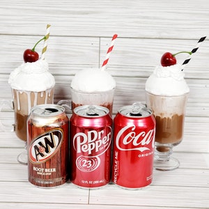 May include: Four 12-ounce cans of soda: A&W Root Beer, Dr Pepper, Coca-Cola, and a fourth can with a red and white label. The cans are in front of four glass mugs filled with a creamy, brown beverage.