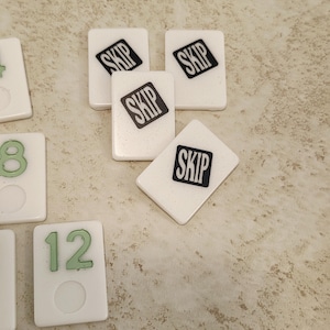 May include: A collection of rectangular game tiles with a white, slightly speckled finish. Three tiles feature the word SKIP in black text on a black background. Other tiles display green numbers, including 8 and 12, with a circular indentation below.