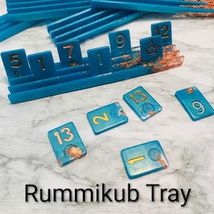 May include: A turquoise Rummikub tray and tiles with gold numbers and accents. The tray holds numbered tiles, and several loose tiles are scattered around. The text "Rummikub Tray" is at the bottom of the image.