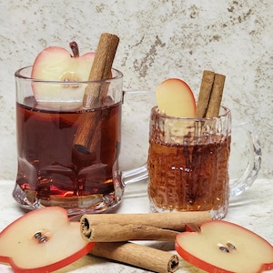 May include: Two clear glass mugs filled with a dark red liquid, garnished with apple slices and cinnamon sticks. The mugs sit on a light-colored surface, with additional apple slices and cinnamon sticks scattered around.