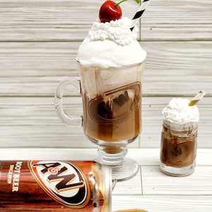 May include: A&W Root Beer themed display featuring a large glass mug and a small glass, both filled with a root beer beverage and topped with whipped cream and a cherry. A spilled can of A&W Root Beer is on its side.