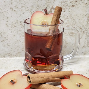May include: A clear glass mug filled with a reddish-brown liquid, an apple slice, and a cinnamon stick. Two additional cinnamon sticks and two apple slices are on the surface in front of the mug. The background is a light, speckled color.