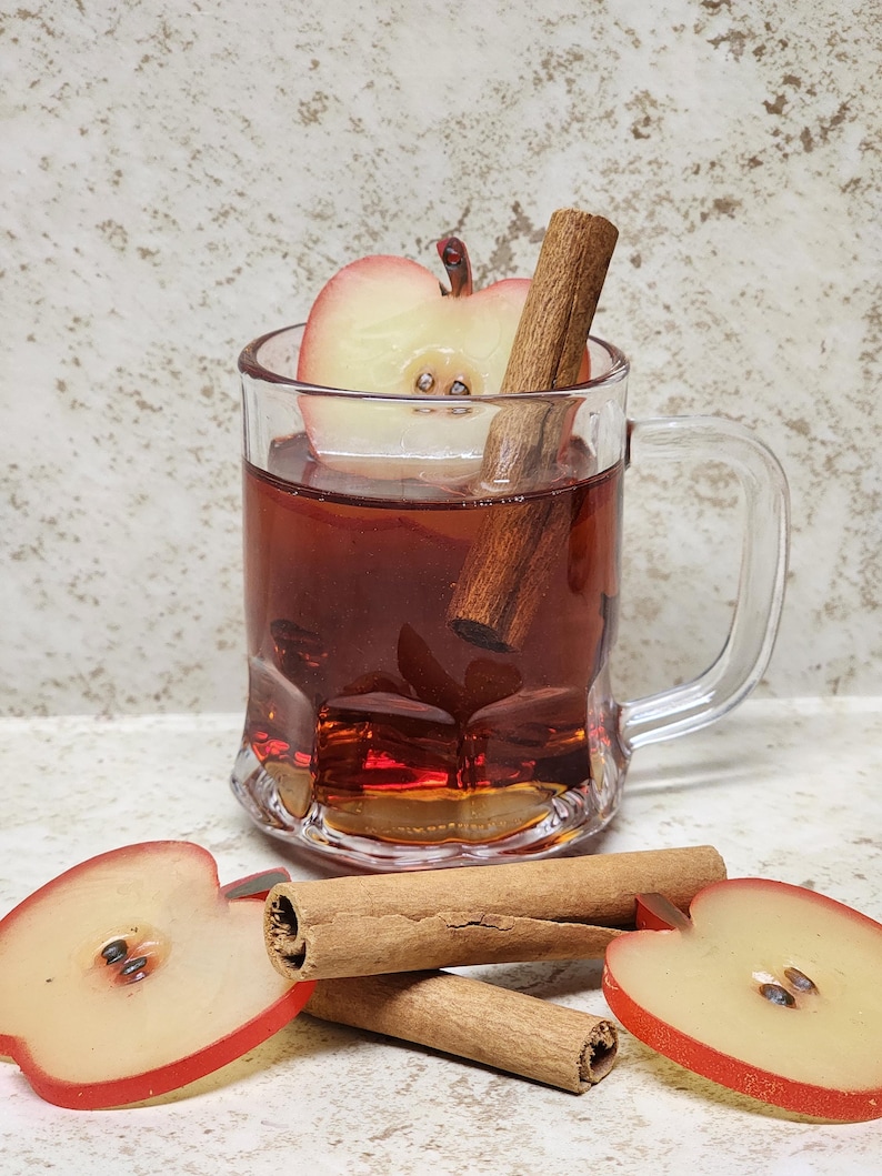 May include: A clear glass mug filled with a reddish-brown liquid, an apple slice, and a cinnamon stick. Two additional cinnamon sticks and two apple slices are on the surface in front of the mug. The background is a light, speckled color.
