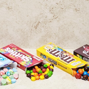 Może przedstawiać: Cztery pudełka cukierków, w tym Sweetarts, Skittles i M&M's. Pudełka są otwarte, a cukierki się z nich wysypują. Pudełko Sweetarts jest różowe, niebieskie i białe. Pudełko Skittles jest czerwone. Pudełka M&M's są żółte i brązowe.