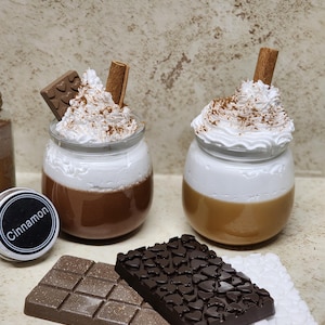 May include: Two glass jars filled with a layered drink, topped with whipped cream, cinnamon, a chocolate square, and a cinnamon stick. Several chocolate bars and a container labeled "Cinnamon" are in the foreground.