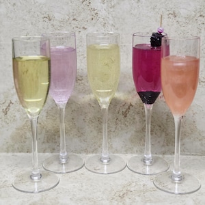 Op de afbeelding: Vijf champagneglazen gevuld met kleurrijke drankjes. De drankjes variëren in kleur van lichtgeel tot roze en paars. Eén glas is gegarneerd met een braambes en een kleine decoratieve prikker. De fluiten zijn van helder glas met lange stelen.