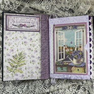 May include: A purple and white scrapbook with a lavender theme. The front cover features a vintage label with the text "Savon a l'Essence de LAVANDE PARIS DEMOLDE". The back cover features a window scene with a watering can filled with lavender flowers.