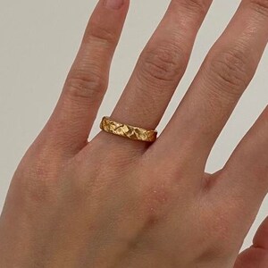 May include: A gold ring with a textured, hammered finish. The ring is worn on the ring finger of a hand.