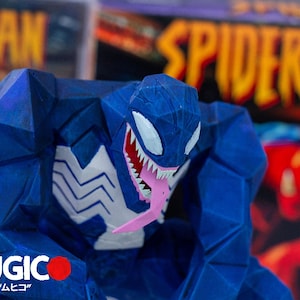 May include: A blue Venom figurine with white detailing and a pink tongue. The figurine has a geometric design, set against a blurred background with the word "SPIDERMAN". The word "MUGIC" is visible at the bottom.