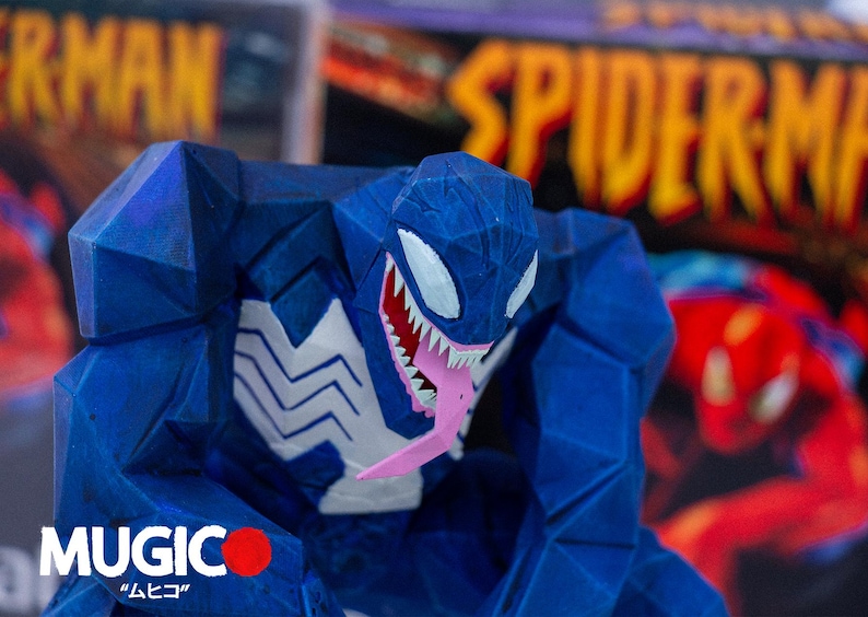 May include: A blue Venom figurine with white detailing and a pink tongue. The figurine has a geometric design, set against a blurred background with the word "SPIDERMAN". The word "MUGIC" is visible at the bottom.
