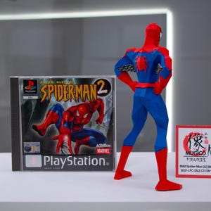 May include: A Spider-Man action figure in a red and blue suit stands next to a PlayStation game case for Spider-Man 2. A small sign with Japanese text and the words "MUGICO FIGURES" is also present.