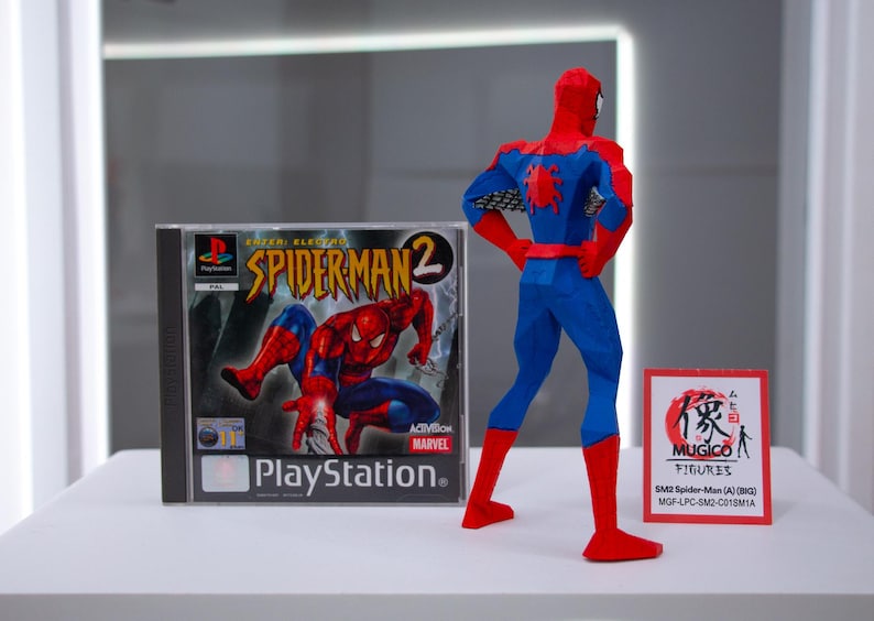 May include: A Spider-Man action figure in a red and blue suit stands next to a PlayStation game case for Spider-Man 2. A small sign with Japanese text and the words "MUGICO FIGURES" is also present.