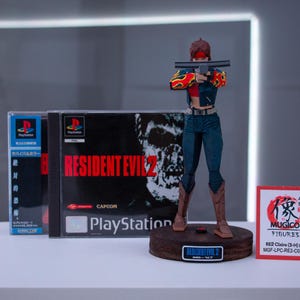 Puede incluir: Una figura de acci&oacute;n roja y negra de una mujer sosteniendo una pistola, de pie sobre una base con las palabras "Resident Evil 2" impresas en ella. La figura es del videojuego Resident Evil 2, y la base es un expositor. La figura lleva una chaqueta roja y negra con un dise&ntilde;o de llamas, pantalones vaqueros azules y botas negras. La figura sostiene una pistola en su mano derecha y la apunta a la c&aacute;mara. La figura est&aacute; de pie sobre una base que est&aacute; hecha de madera y tiene una base negra. La base tiene las palabras "Resident Evil 2" impresas en ella en letras blancas. La figura es del videojuego Resident Evil 2, que fue lanzado para PlayStation en 1998.