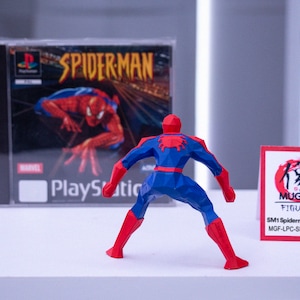 May include: A red, white, and blue Spider-Man action figure stands before a PlayStation game case. The game case displays "SPIDERMAN" and the Marvel logo. A small card with Japanese text and "MUGICO FIGURES" is also present.