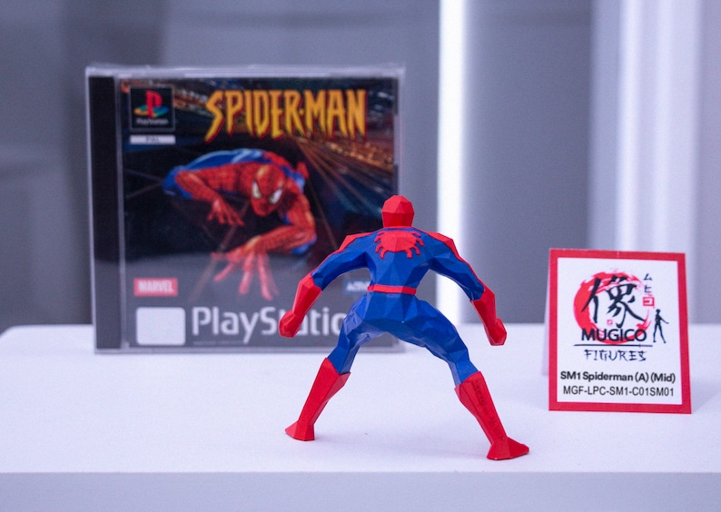 May include: A red, white, and blue Spider-Man action figure stands before a PlayStation game case. The game case displays "SPIDERMAN" and the Marvel logo. A small card with Japanese text and "MUGICO FIGURES" is also present.
