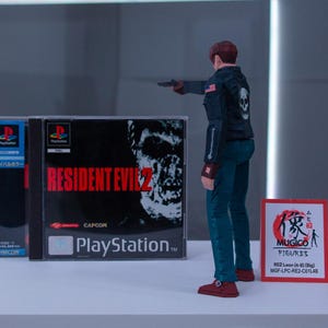 May include: A video game case for Resident Evil 2 for the Playstation console. The case is black with red text and a zombie on the cover. The case is next to a small action figure of a man in a black leather jacket and blue jeans holding a gun.