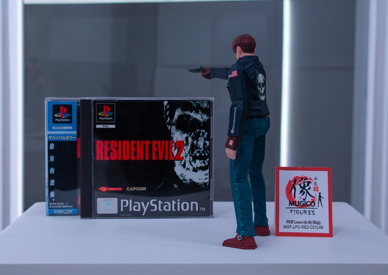 May include: A video game case for Resident Evil 2 for the Playstation console. The case is black with red text and a zombie on the cover. The case is next to a small action figure of a man in a black leather jacket and blue jeans holding a gun.