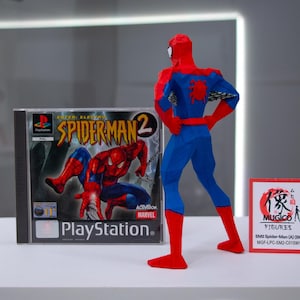 May include: A blue and red Spider-Man action figure stands next to a PlayStation game case for Spider-Man 2. The game case features the Spider-Man logo and character art. A small red sign with Japanese text is also present.