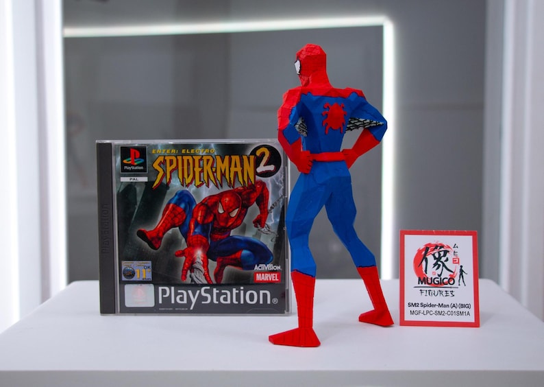 May include: A blue and red Spider-Man action figure stands next to a PlayStation game case for Spider-Man 2. The game case features the Spider-Man logo and character art. A small red sign with Japanese text is also present.