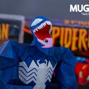 Puede incluir: Una figura azul de Venom con un emblema de ara&ntilde;a blanco en el pecho y dientes blancos. La figura tiene una boca roja y una lengua rosa. El fondo incluye portadas de c&oacute;mics con las palabras "Spider-Man" y "MUGIC" en blanco y caracteres japoneses.