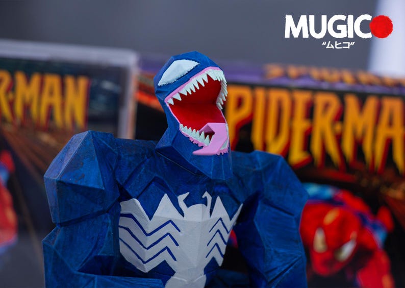 Puede incluir: Una figura azul de Venom con un emblema de ara&ntilde;a blanco en el pecho y dientes blancos. La figura tiene una boca roja y una lengua rosa. El fondo incluye portadas de c&oacute;mics con las palabras "Spider-Man" y "MUGIC" en blanco y caracteres japoneses.
