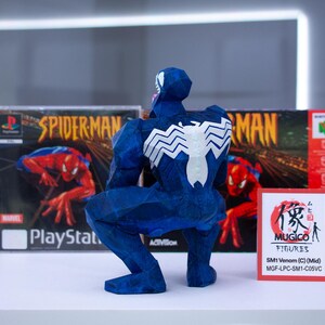 Puede incluir: Una figura azul de Venom con un emblema de ara&ntilde;a blanco en la espalda, posando frente a una caja de juego de Spider-Man para PlayStation y una caja de Nintendo 64. Un peque&ntilde;o letrero rojo dice "MUGICO FIGURES".