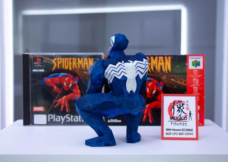 Puede incluir: Una figura azul de Venom con un emblema de ara&ntilde;a blanco en la espalda, posando frente a una caja de juego de Spider-Man para PlayStation y una caja de Nintendo 64. Un peque&ntilde;o letrero rojo dice "MUGICO FIGURES".