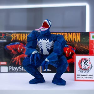 Puede incluir: Una figura azul de Venom con un emblema de ara&ntilde;a blanco, boca abierta y lengua larga, posando frente a una caja de juego de Spider-Man PlayStation y una caja roja. Es un objeto de colecci&oacute;n.