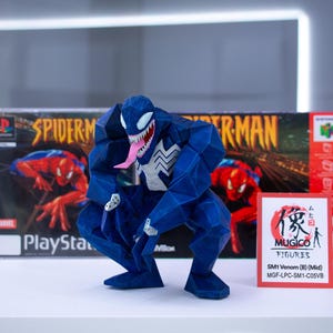 May include: A blue geometric Venom figure with a white spider symbol and pink tongue, posed in front of a Spider-Man PlayStation game box. A small card with Japanese text and "MUGICO FIGURES" is also present.