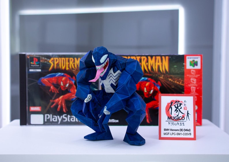 May include: A blue geometric Venom figure with a white spider symbol and pink tongue, posed in front of a Spider-Man PlayStation game box. A small card with Japanese text and "MUGICO FIGURES" is also present.