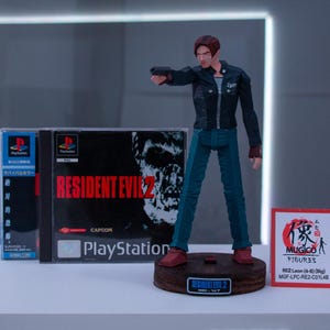 May include: A plastic action figure of Leon S. Kennedy from the Resident Evil 2 video game. The figure is holding a gun and is wearing a black jacket, blue jeans, and brown boots. The figure is standing on a wooden base with the words "Resident Evil 2" printed on it. The figure is next to a PlayStation game case for Resident Evil 2. The game case is blue and white with the words "Resident Evil 2" printed in red. The game case is next to a small white card with the words "Mugico Figures" printed on it.
