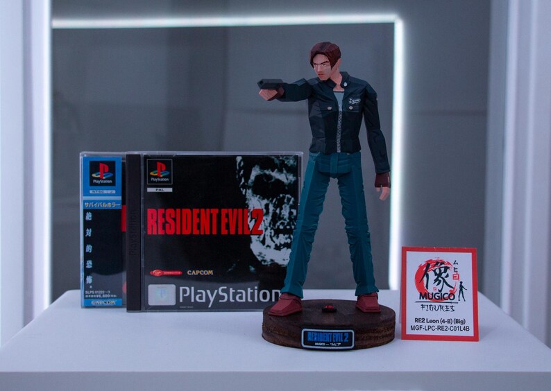 May include: A plastic action figure of Leon S. Kennedy from the Resident Evil 2 video game. The figure is holding a gun and is wearing a black jacket, blue jeans, and brown boots. The figure is standing on a wooden base with the words "Resident Evil 2" printed on it. The figure is next to a PlayStation game case for Resident Evil 2. The game case is blue and white with the words "Resident Evil 2" printed in red. The game case is next to a small white card with the words "Mugico Figures" printed on it.