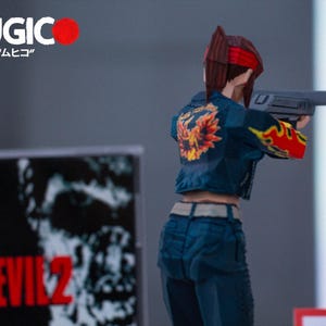 Puede incluir: Una figura de acci&oacute;n azul y roja de una mujer sosteniendo una pistola. La figura lleva una chaqueta vaquera azul con un dise&ntilde;o de llamas amarillas y naranjas en la espalda. La figura est&aacute; de pie frente a una caja en blanco y negro con el texto "Resident Evil 2" en ella.