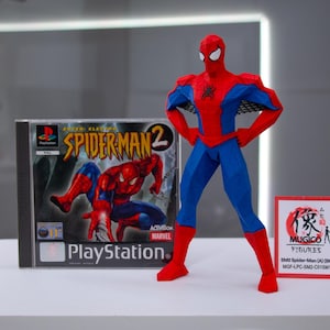 May include: A red, blue, and black Spider-Man action figure stands next to a PlayStation game case for Spider-Man 2. The game case features the Spider-Man 2 logo and a graphic of Spider-Man. A small sign with Japanese text is also present.