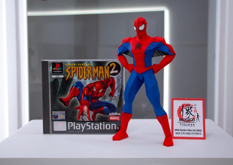 May include: A red, blue, and black Spider-Man action figure stands next to a PlayStation game case for Spider-Man 2. The game case features the Spider-Man 2 logo and a graphic of Spider-Man. A small sign with Japanese text is also present.
