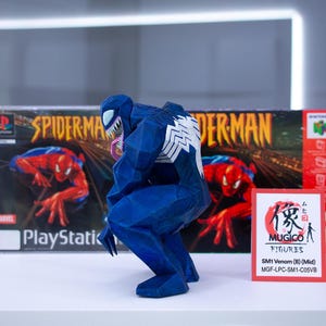 May include: A blue Venom figurine with white detailing, in a crouching pose. It's positioned in front of a PlayStation game box and a Nintendo 64 game box, both with Spider-Man artwork. A small card with Japanese text and the Mugico Figures logo is also visible.