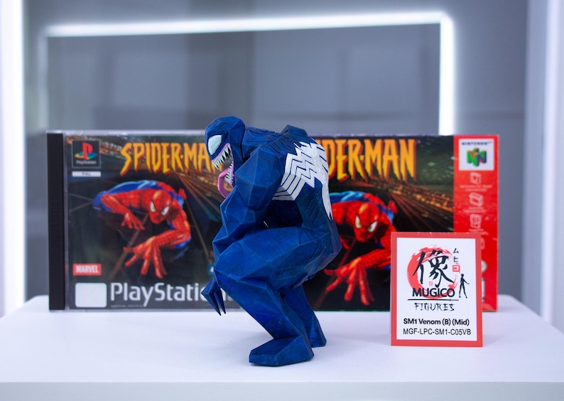 May include: A blue Venom figurine with white detailing, in a crouching pose. It's positioned in front of a PlayStation game box and a Nintendo 64 game box, both with Spider-Man artwork. A small card with Japanese text and the Mugico Figures logo is also visible.