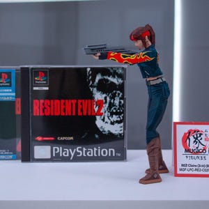 Puede incluir: Una figura de acci&oacute;n roja y negra de una mujer sosteniendo una pistola, de pie frente a dos cajas de juegos de PlayStation para Resident Evil 2. Las cajas de juegos est&aacute;n en japon&eacute;s e ingl&eacute;s. La figura de acci&oacute;n est&aacute; etiquetada "MUGICO FIGURES RE2 Claire (3-4) (Big) MGF-LPC-RE2-C02C3H".