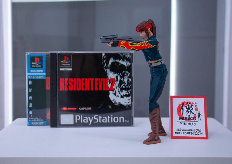 Puede incluir: Una figura de acci&oacute;n roja y negra de una mujer sosteniendo una pistola, de pie frente a dos cajas de juegos de PlayStation para Resident Evil 2. Las cajas de juegos est&aacute;n en japon&eacute;s e ingl&eacute;s. La figura de acci&oacute;n est&aacute; etiquetada "MUGICO FIGURES RE2 Claire (3-4) (Big) MGF-LPC-RE2-C02C3H".