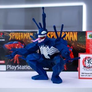 May include: A blue Venom figurine with a white spider emblem, pink tongue, and sharp teeth, posed in front of a PlayStation Spider-Man game case and a Nintendo 64 game box. A small sign reads "MUGICO FIGURES".