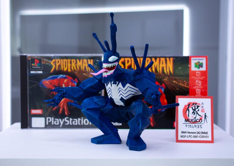 May include: A blue Venom figurine with a white spider emblem, pink tongue, and sharp teeth, posed in front of a PlayStation Spider-Man game case and a Nintendo 64 game box. A small sign reads "MUGICO FIGURES".