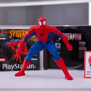 May include: A red and blue Spiderman action figure in a dynamic pose, standing in front of two Playstation game cases. The figure is made of a low-poly style, with a black spider logo on the chest. The game cases are for the Spiderman game for the Playstation console.