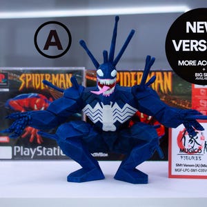 May include: A dark blue Venom figurine with white accents and a pink tongue, posed in front of a Spider-Man PlayStation game case. The figure has a jagged design and the text "NEW VERSION!" is visible.