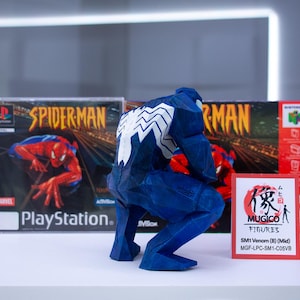 May include: A blue Venom figurine with white detailing, positioned before two Spider-Man video game boxes. The PlayStation and Nintendo 64 game boxes are visible. A small card with Japanese text is in front of the figurine.