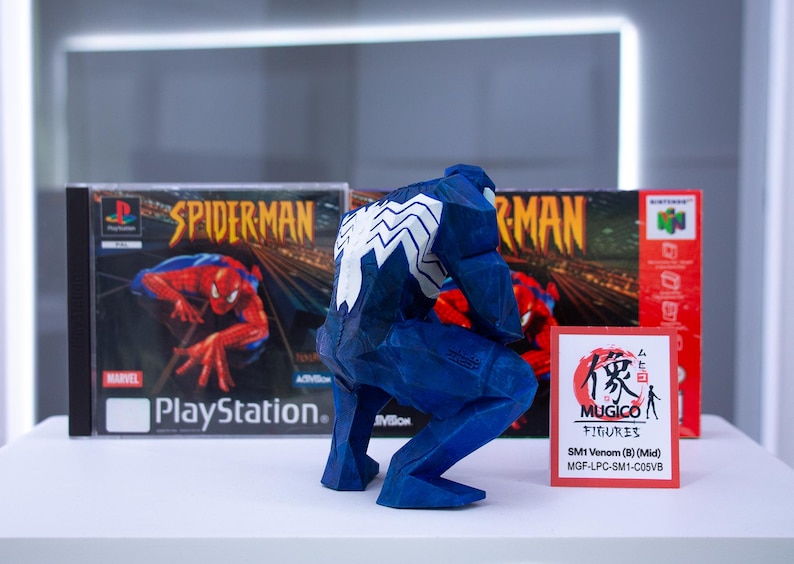 May include: A blue Venom figurine with white detailing, positioned before two Spider-Man video game boxes. The PlayStation and Nintendo 64 game boxes are visible. A small card with Japanese text is in front of the figurine.