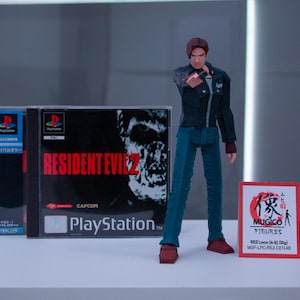 May include: A video game case for Resident Evil 2 for the Playstation console. The case is black with red text and a zombie on the cover. A red and white action figure of Leon S. Kennedy from the game is standing in front of the case. The action figure is holding a gun and has a serious expression on his face. A red and white card with Japanese text and the words "MUGICO FIGURES" is in the foreground.
